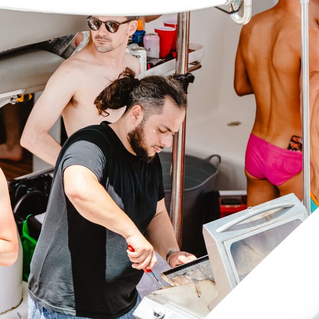 A man with a beard and hair in a bun wearing a black shirt is cooking on a grill. Surrounding him are several shirtless individuals, some sporting brightly colored swimwear, standing and socializing on a boat. The sun shines down as laughter fills the air, creating a lively atmosphere.