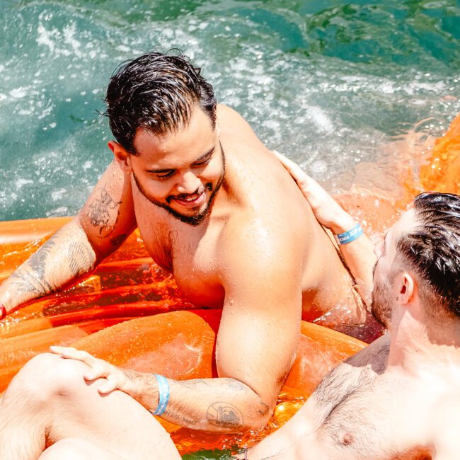 Two men are relaxing on an orange float in the water, smiling at one another. Both are shirtless and appear to be enjoying a sunny day. One is leaning back with sunglasses in hand while the water splashes around them. An inflatable beach ball floats nearby, adding to the scene of summer fun.