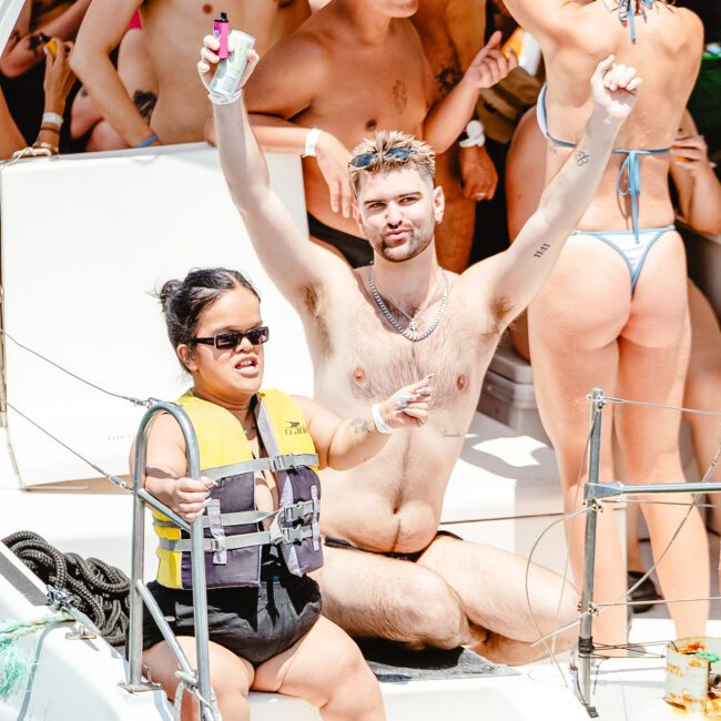 A group of people are enjoying a sunny day on a boat. They are in swimwear, and one person in the center is raising their arms with enthusiasm. Others around them are engaging in conversation and enjoying beverages. There is a well-stocked lifeboat and safety equipment on board for everyone’s peace of mind.