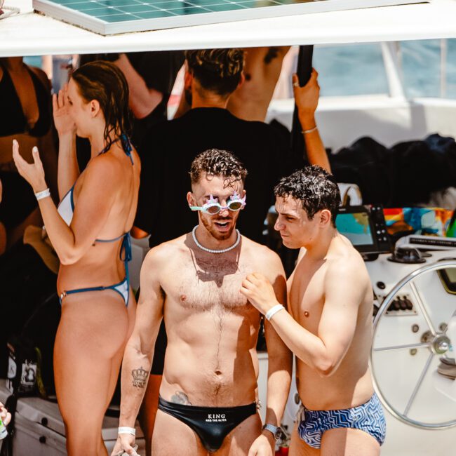 A group of people are enjoying a sunny day on a yacht. In the foreground, two men in swimwear are standing; one wearing sunglasses and a necklace, while the other seems to be talking to him. Others are in the background with towels and equipment visible, savoring the tranquility of the open sea.