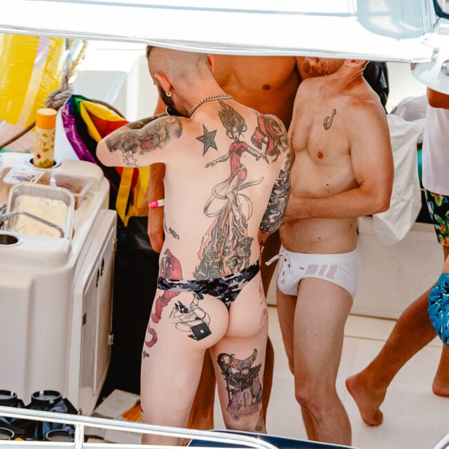 Several people are on a boat, some with visible tattoos and colorful swimwear. They appear to be socializing and enjoying a sunny day. One person stands out with extensive body tattoos and black swim trunks. Logos and small decorations, including nautical elements, are seen in the background.