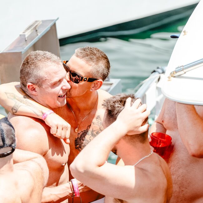 A group of men are at a party on a boat, basking in the festive atmosphere. Two shirtless men in the center are embracing, one kissing the other's cheek while others hold red cups. The sun is shining brightly above them.