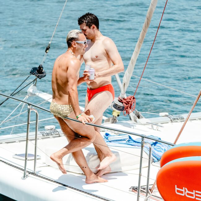 Two men in swimsuits stand on a sailboat: one in a yellow and brown swimsuit and the other in a red swimsuit. The boat floats on clear blue water, with a paddleboard nearby. They appear relaxed, enjoying their time under the bright sunlight, with mountain views enhancing the serene atmosphere.