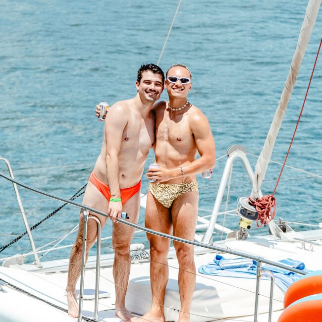 Two men in swimwear stand on the deck of a boat, smiling and embracing. The man on the left wears red swim trunks, while the man on the right sports yellow swim trunks and sunglasses. They are surrounded by water, enjoying a sunny day.