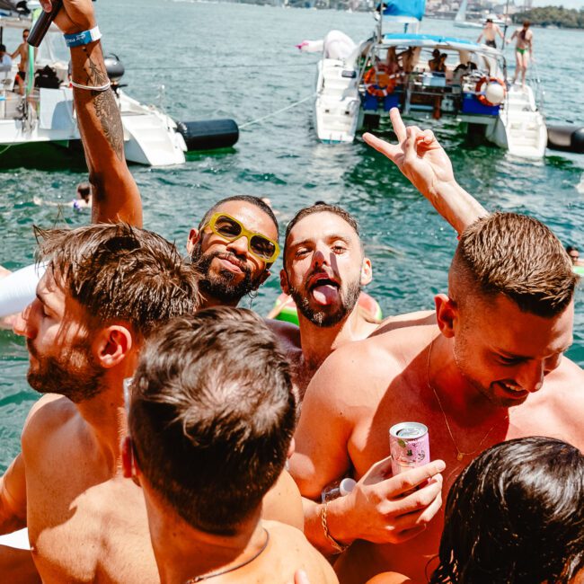 A group of shirtless men having fun on a boat, some holding drinks and smiling for the camera. In the background, other people are enjoying boats and water activities, with a stunning city skyline in the distance. It appears to be a sunny day filled with joy.