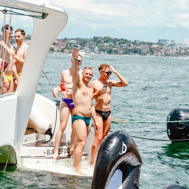 A group of people in swimwear are gathered on and around a boat, enjoying a sunny day on the water. One person is waving at the camera while others are smiling and having fun. The backdrop features a stunning cityscape with buildings and greenery visible across the shimmering water.