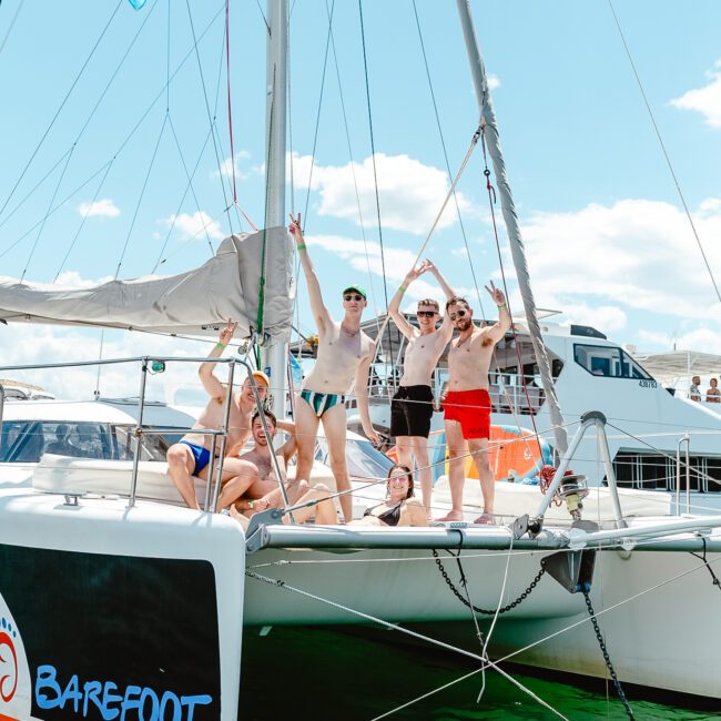 A group of six people in swimsuits are posing and smiling on the front deck of a catamaran named "Barefoot" on a sunny day. Some are making peace signs with their hands. The catamaran is gently sailing on clear water, with a few clouds in the background sky.