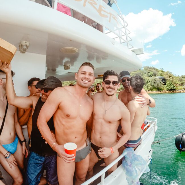 A group of people on a boat, many in swimwear and holding drinks, are enjoying a sunny day. The background showcases water and greenery. Two shirtless men in the foreground are smiling and posing for the camera, one holding a red cup and the other, a yellow drink cup. The vibrant setting radiates joy.