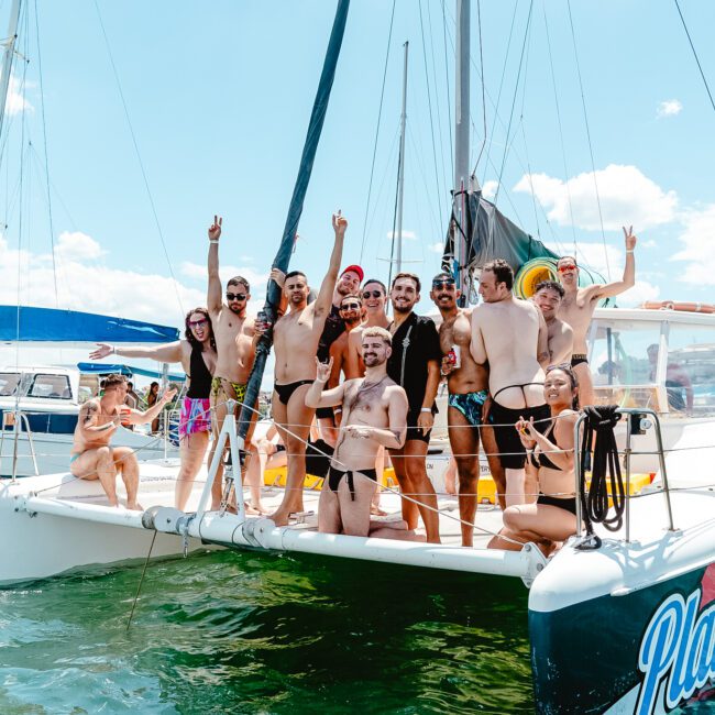 A group of people in swimwear stand and sit on a catamaran sailboat in a sunny harbor, many posing and smiling for the camera, some making peace signs. The boat is stationary amid bright blue skies and other boats with colorful sails in the background.