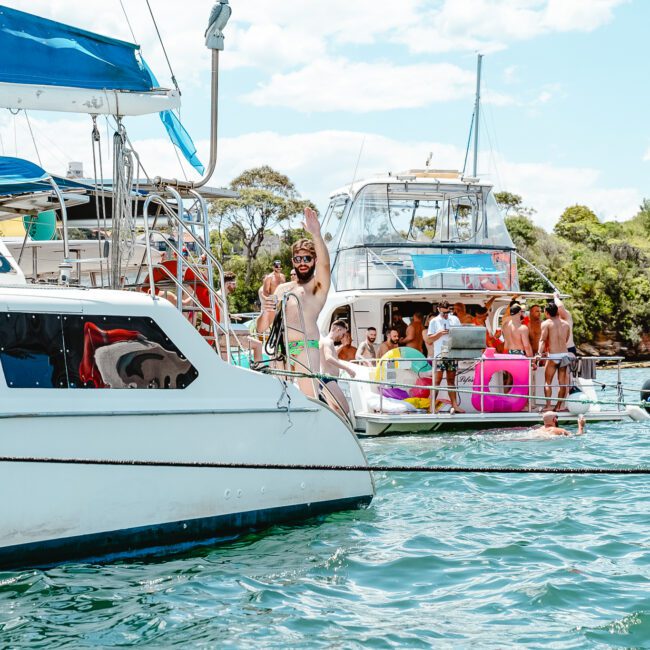 A group of people enjoying a sunny day on several anchored boats near the shore. Some are standing on the decks, while one person stands on a floating platform between boats. Bright inflatables and swimwear add splashes of color to the scene, with trees and a clear blue sky in the background.