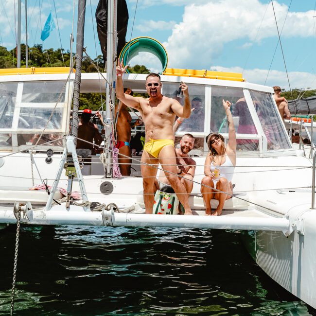 A group of people on a sailboat enjoying a sunny day, with one man in yellow swimwear confidently holding a drink at the front. Others sit and relax behind him, all in swimwear, smiling and celebrating together. The water sparkles under the clear blue sky.