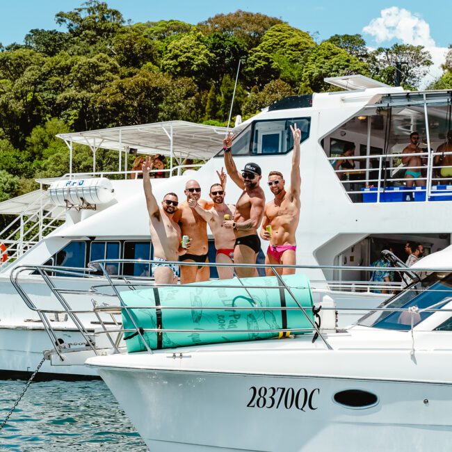 A group of six men in swimwear stand on the deck of a boat, posing and cheering towards the camera. Another boat is visible in the background. The scene is set on a sunny day with lush greenery and crystal-clear water in the distance.