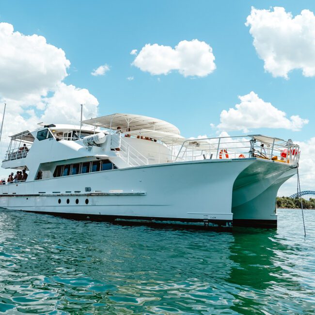 A large white yacht floating on calm water under a clear blue sky with scattered clouds. People can be seen on the upper and lower decks of the yacht. In the background, a distant bridge spans across a picturesque body of land adorned with lush greenery.