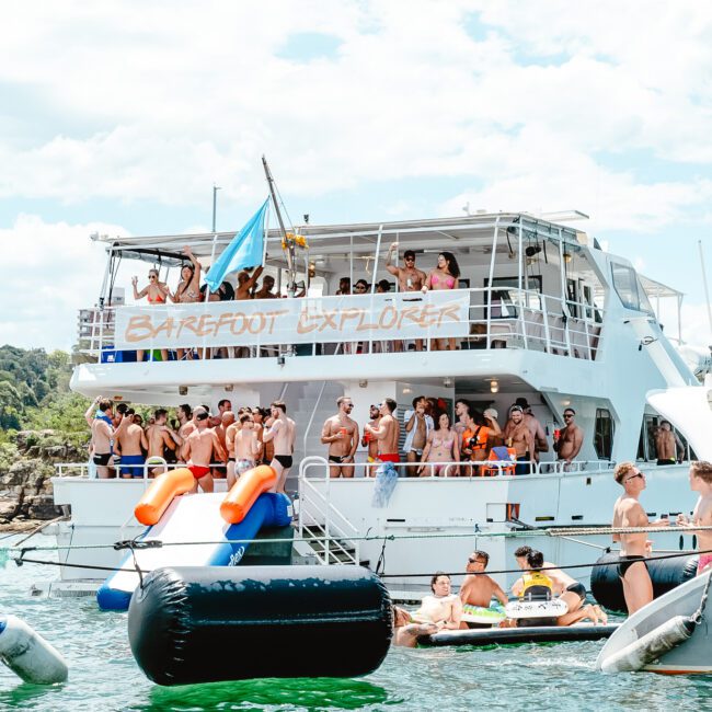 A large boat named "BAREFOOT EXPLORER" is surrounded by inflatable rafts teeming with people who are either sunbathing or socializing. A blue flag flutters on the boat, adding to the festive and vibrant atmosphere on this sunny day.