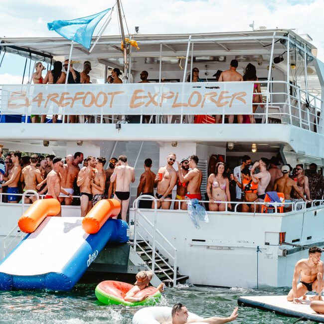 The vibrant boat named "Barefoot Explorer" is hosting a lively party with many people in swimsuits. Two inflatable slides are attached to the boat, leading to cheerful swimmers in the water, some on floats. The backdrop features lush trees and a city skyline under a pristine blue sky.