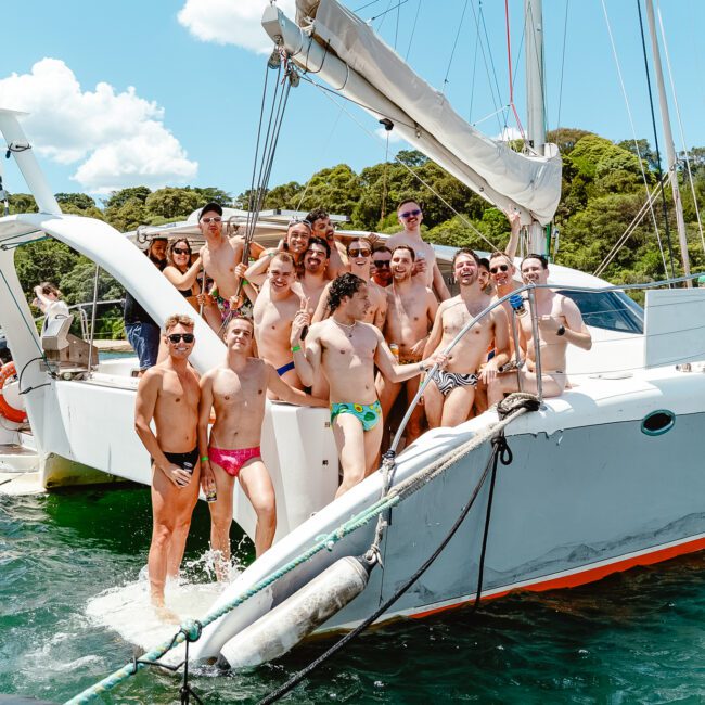 A large group of people pose together on a sailboat in a sunny location. Most are wearing swimsuits, and they appear to be enjoying the day. The backdrop includes a forested coast and blue water. There are logos for GAYM and Yacht Social Club, highlighting their collaboration for this vibrant event.