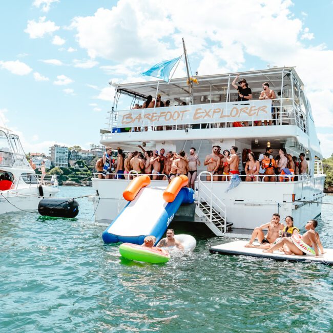 A large, multi-story boat named "Barefoot Explorer" is anchored on a sunny day, teeming with people in swimsuits. The boat boasts a water slide, and some are enjoying inflatables in the water. Other boats and lush greenery form a picturesque background.