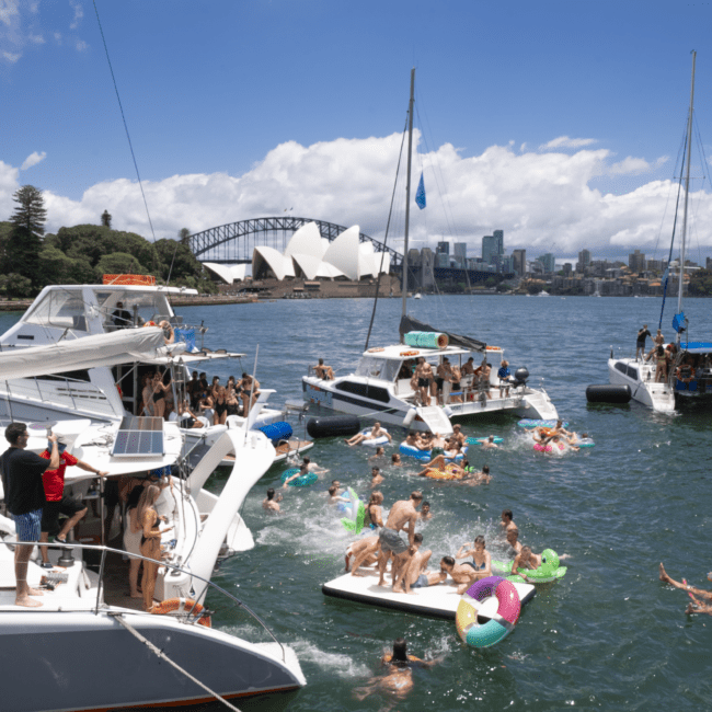 A lively scene of people enjoying a sunny day on and around several boats in a harbor, with inflatables floating in the water. The iconic Sydney Opera House and Sydney Harbour Bridge are visible in the background under a partly cloudy sky, capturing the vibrant atmosphere of the city.