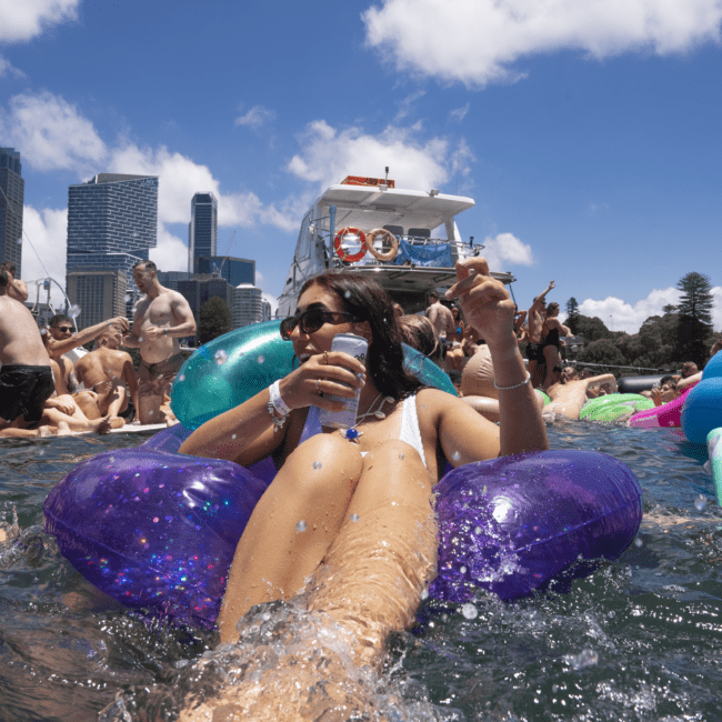 A lively group of people enjoys a sunny day on the water, some on inflatable floats with drinks in hand, others standing nearby. Boats and skyscrapers are visible in the background. The clear sky adds to the vibrant, festive atmosphere enhanced by cheerful laughter.