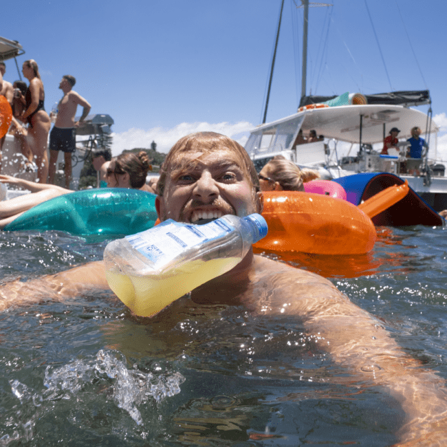 A man in a body of water bites onto a bottle, surrounded by people on vibrant inflatables. In the background, a white boat with more people can be seen under a clear blue sky. The scene is lively and festive, suggesting a joyful gathering on the water.