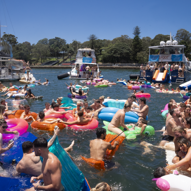 A large group of people is enjoying a sunny day on the water, swimming and floating on colorful inflatables. Paddleboards are scattered around with more people on them. Boats are anchored nearby. Trees and a grassy area can be seen in the background. The atmosphere is lively and festive.