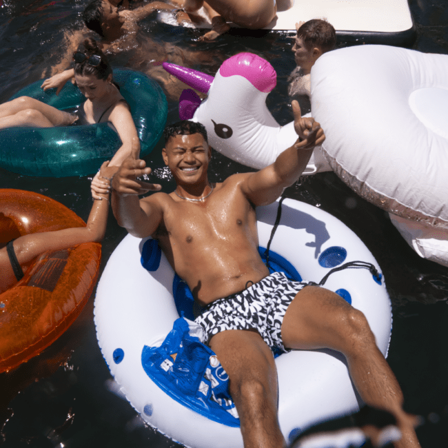 A man in patterned swim trunks smiles and gives two thumbs up while lounging on an inflatable ring in a pool. Surrounding him, people are enjoying the water on various inflatable floats, including a colorful unicorn. The scene is lively and sunlit with everyone having fun under the blue sky.