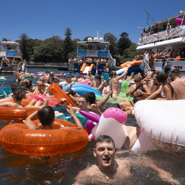 A lively scene of people enjoying a sunny day on the water, floating on colorful inflatables near boats. Some are on a unicorn float, while others are on orange and donut-shaped ones. The gentle breeze rustles the trees, and clear skies frame the background as music and laughter fill the air.