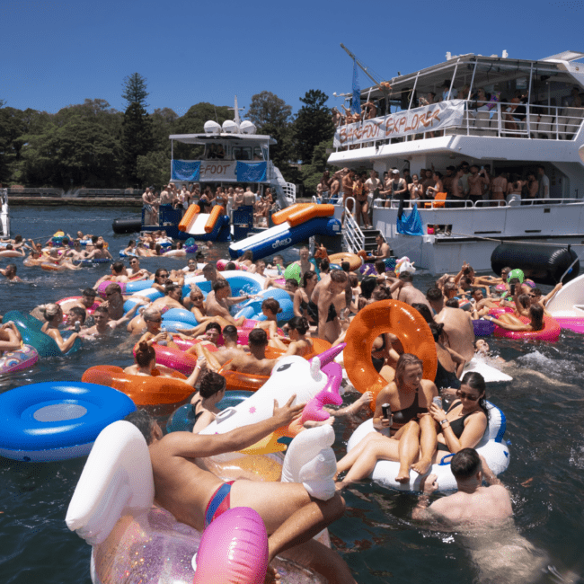 A lively scene of people enjoying a sunny day on the water with numerous inflatable pool floats, including unicorns and doughnuts. A large boat is docked nearby. Some are diving into the sparkling water, while others relax under colorful canopies. The atmosphere is festive and energetic.