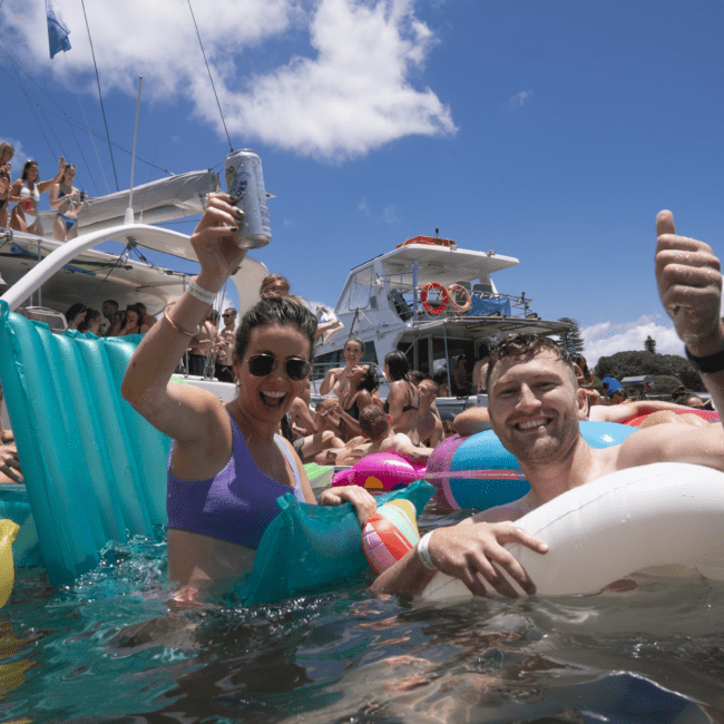 A woman and man are smiling and holding drinks while floating in the water at a lively boat party. Several others are socializing and relaxing on inflatable rafts and nearby boats under a sunny, partly cloudy sky.