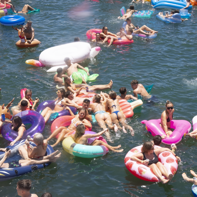 A large group of people enjoying a sunny day on the water, floating on various colorful inflatable rafts like unicorns, donuts, and tubes. Boats are visible in the background with more people relaxing and socializing. Some are even playing music to enhance the lively atmosphere.