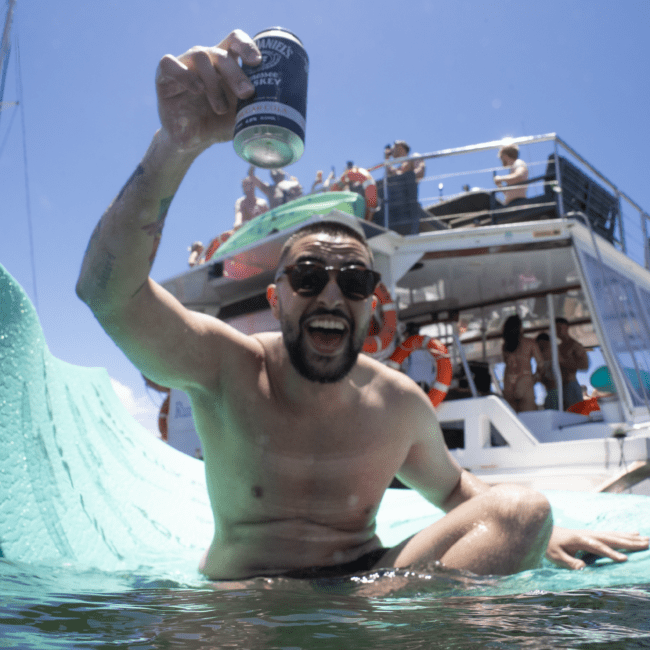 A shirtless person wearing sunglasses holds up a canned drink and smiles widely while sitting on a float in the water. In the background, there is a red double-decker boat with several people on board under a clear blue sky.