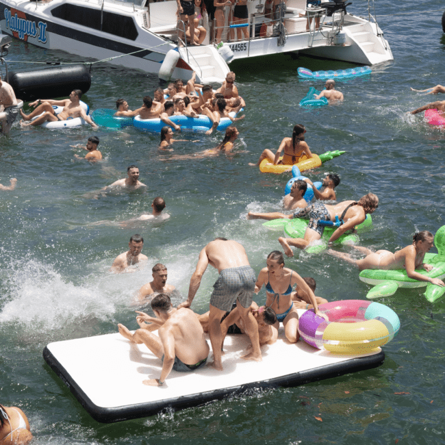 A crowded lake scene with people swimming, floating on inflatable rafts, and playing on a large floating mat. In the background, a boat named "Papayos II" is anchored, with more people on board. Everyone appears to be enjoying a fun-filled, sunny day on the water.