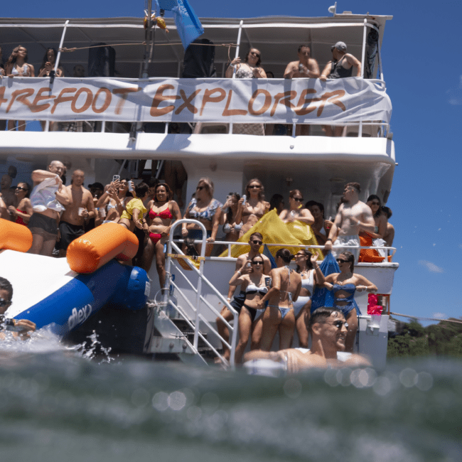 A group of people enjoying a sunny day on a two-story boat named "Barefoot Explorers." Some relax on the deck, while others use an inflatable slide to plunge into the serene water. The sky is clear with a few scattered clouds, adding to the picturesque scene.