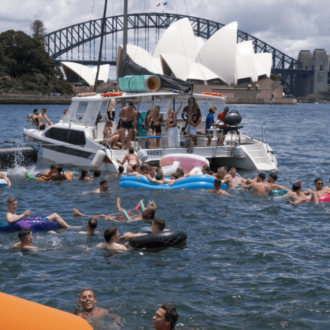 A group of people enjoy a sunny day on a boat and in the water, with some floating on inflatable tubes. The iconic Sydney Opera House and Harbour Bridge are visible in the background, perfectly capturing a lively aquatic scene.