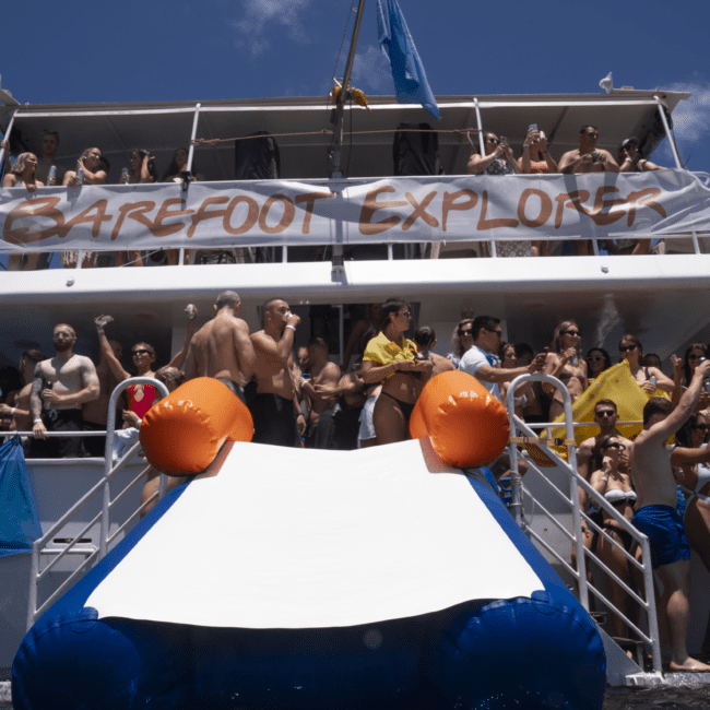 A large group of people in swimsuits are enjoying themselves on a boat, named "Barefoot Explorer". The boat has multiple decks with people on each level. The lower deck includes a fun slide leading to the water, and the sky is clear and sunny—a perfect day for adventure!