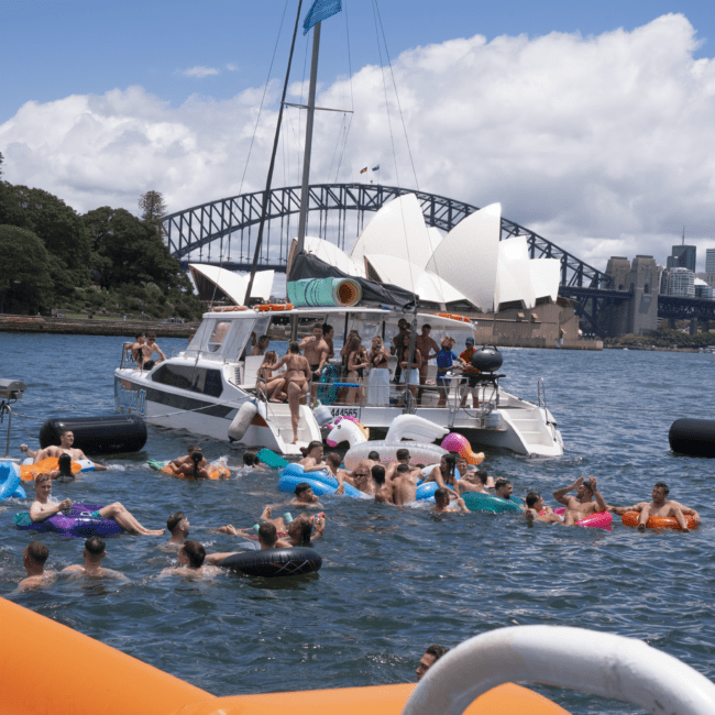 A lively scene of people swimming and floating on inflatable tubes near a yacht party. The yacht is anchored with several people on deck enjoying the day. The iconic Sydney Opera House and Harbour Bridge are visible in the background under a sunny sky, creating a picturesque setting.