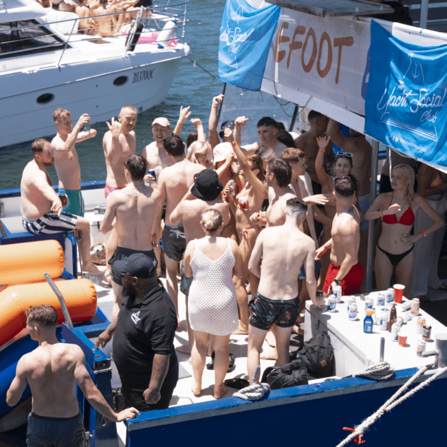 A lively group of people in swimsuits enjoy a party on a boat, with drinks and snacks. Another boat with more people is docked nearby. A blue banner with "8FOOT" flutters in the breeze. The sunny scene is festive, with attendees dancing and socializing, creating an energetic atmosphere.