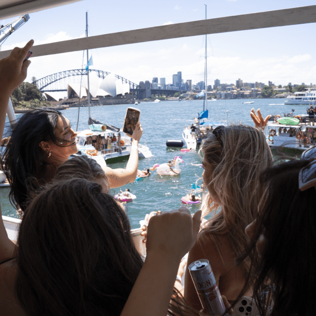 A group of people aboard a boat celebrate, many holding drinks. Some capture the moment with phones while facing the iconic Sydney Opera House and Harbour Bridge in the background. Other boats and floating toys add to the lively atmosphere under a clear blue sky, adorned with fluffy clouds.