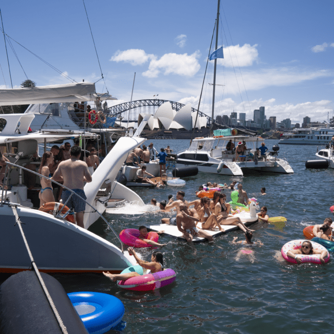 A lively scene of people enjoying a sunny day on the water. A group is gathered on yachts and in the water on colorful inflatable floats. In the background, a picturesque bridge and city skyline are visible under a bright blue sky with scattered clouds.