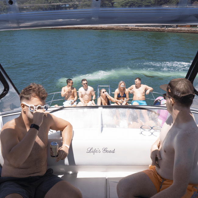 A group of young adults enjoy a sunny day on a boat and floating dock on a lake. Two men in the foreground are sitting on the boat's back bench, while others lounge with drinks. A dog frolics nearby as the green shoreline and clear water extend into the distance.