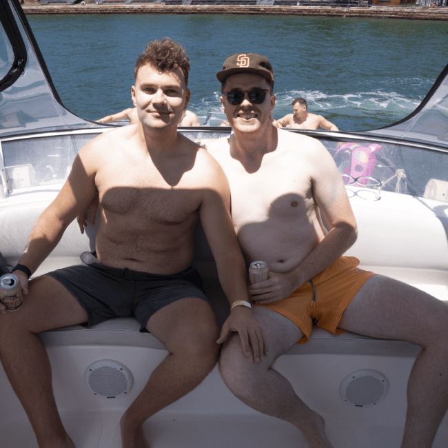 Two men are sitting on the edge of a boat, smiling at the camera. They are shirtless and holding canned drinks. One is wearing orange swim shorts, a dark cap, and sunglasses. Trees and water create a serene backdrop, adding to their relaxed vibe.