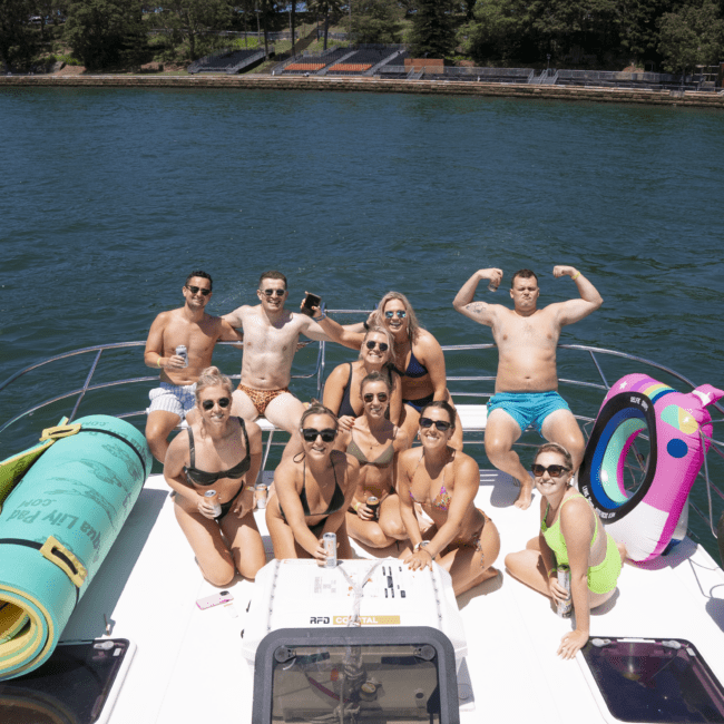 A group of ten people are gathered on the deck of a boat on a sunny day. Some are sitting and others are standing, with one man flexing his muscles. They are surrounded by water with a forested shoreline in the background. Colorful pool floats dot the deck amid the cheerful scene.