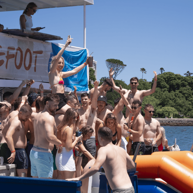 A group of people in swimsuits enjoy a lively party on a boat under sunny skies. Some are dancing, waving, and chatting. The boat features a sign with the word "Barefoot." Vibrant green foliage and trees provide a beautiful backdrop.