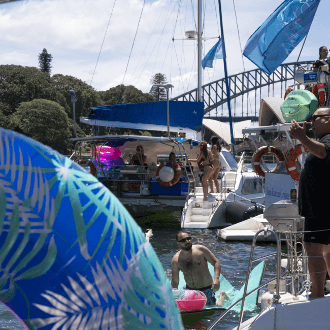 People are enjoying a sunny day on boats decorated with colorful floatation devices. In the background, a bridge is visible amidst trees, adding to the scenic view. Some individuals are swimming and engaging in activities, while others are relaxing on the boats in vibrant swimwear.