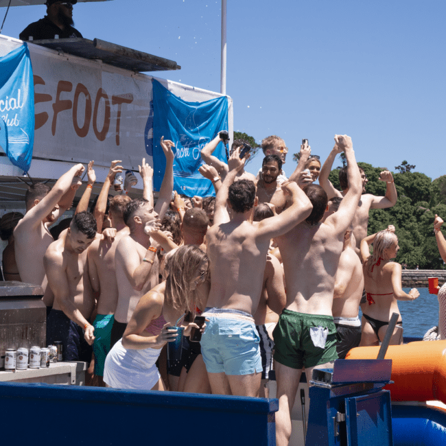 A group of people in swimsuits are gathered on a boat, joyfully dancing and celebrating under a clear blue sky. They hold drinks, surrounded by banners and towels. The vibrant scene captures the lively and energetic atmosphere of a summer yacht party.