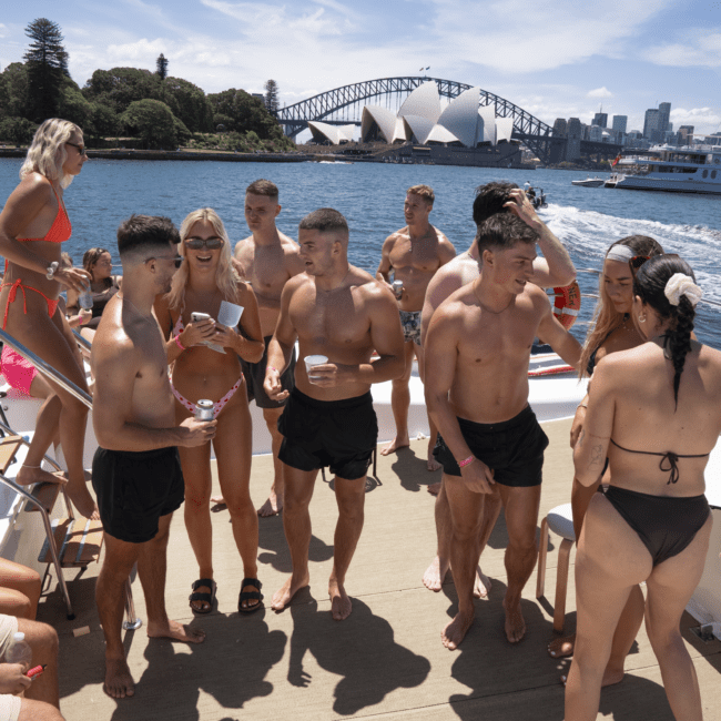 A group of people in swimsuits are gathered on a boat deck overlooking a body of water with the Sydney Opera House and Sydney Harbour Bridge in the background. Some are standing and talking, while others are looking out at the scenic view. It is a sunny day.