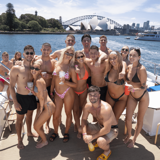 A group of people in swimwear pose together on a boat in a sunny location. They are smiling and holding drinks, with the Sydney Opera House and Sydney Harbour Bridge visible in the background, along with the city skyline and scenic trees.