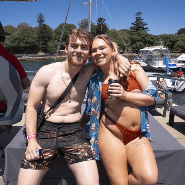 Two smiling people are sitting on a boat dock. The person on the left is shirtless, wearing black swim trunks and a shoulder bag, while the person on the right sports an orange bikini and a blue cover-up. Both have one arm around each other, with boats and trees providing a serene background.

Keyword used: serene