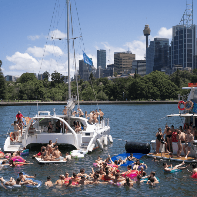 A lively scene on a sunny day with numerous people partying on a catamaran and an inflatable boat in the water. Many are swimming and floating on inflatables. The city skyline, featuring a prominent iconic tower, can be seen in the background.