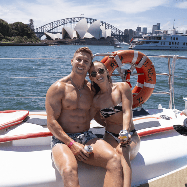 A shirtless man and a woman in a bikini sit together on the edge of a boat, smiling at the camera. The backdrop features the stunning Sydney Opera House, the Harbour Bridge, and several boats on the sparkling water. The woman enjoys her drink on this sunny day, epitomizing a perfect summer moment.